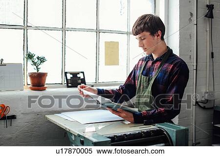 Stock Photography - Young craftsman by window, leaning on letterpress machine and looking at clipboard in book arts studio. Fotosearch