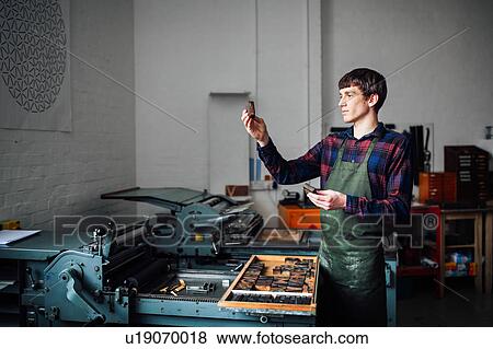 Stock Photo - Young craftsman next to letterpress machine, holding up letter in print workshop. Fotosearch