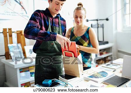 Young craftsman with customer, placing book into bag in arts bookshop View Large Photo Image Stock Image - Young craftsman with customer, placing book into bag in arts bookshop. Fotosearch