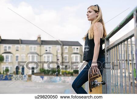 Stock Image - Young female skateboarder leaning against fence in skatepark. Fotosearch