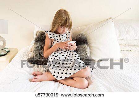 Stock Photo - Young girl sitting on bed playing on an electronic device. Fotosearch