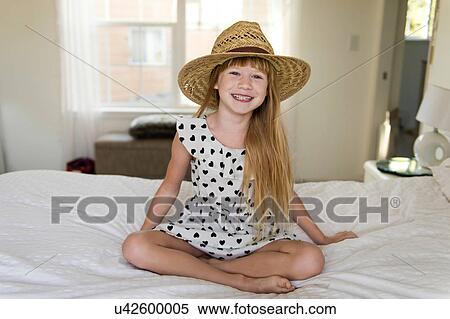 Stock Photography - Young girl sitting on bed smiling, wearing straw hat. Fotosearch