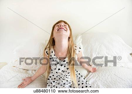 Stock Image - Young girl sitting on bed throwing head back. Fotosearch