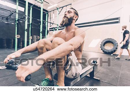 Picture - Young male cross trainer training on rowing machine in gym. Fotosearch