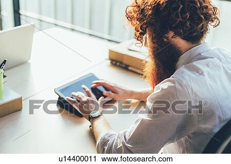 Stock Image - Young male hipster with red hair and beard using digital tablet touchscreen at desk. Fotosearch