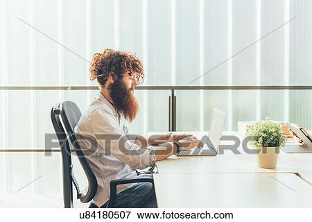 Stock Photo - Young male hipster with red hair and beard typing on laptop at desk. Fotosearch