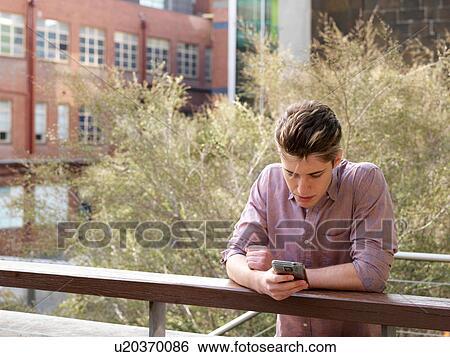 Stock Photograph - Young man leaning on fence, using smartphone, outdoors. Fotosearch