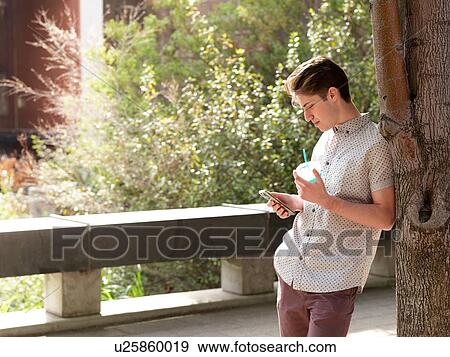 Young man outdoors, leaning against tree, using smart phone, holding takeaway cup View Large Photo Image Stock Photo - Young man outdoors, leaning against tree, using smart phone, holding takeaway cup. Fotosearch