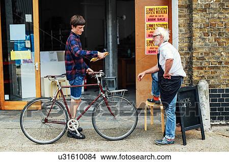 Picture - Young man with pushbike looking at post and standing with senior craftsman outside print workshop. Fotosearch