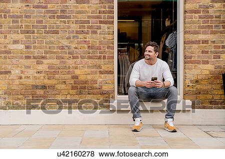 Stock Photo - Young man with smartphone sitting on doorstep, Kings Road, London, UK. Fotosearch