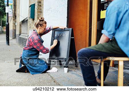 Stock Image - Young woman kneeling on pavement and writing on blackboard outside workshop. Fotosearch