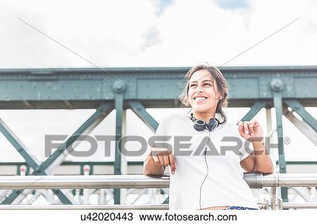 Stock Image - Young woman standing on bridge, headphones around neck, smiling. Fotosearch