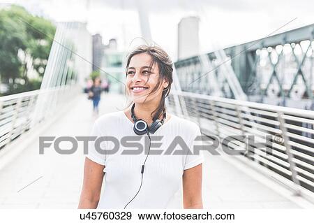 Stock Photo - Young woman standing on bridge, headphones around neck, smiling. Fotosearch