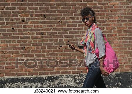Stock Image - Young woman walking against brick wall. Fotosearch