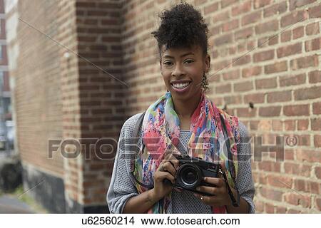 Picture - Young woman with digital camera against brick wall. Fotosearch