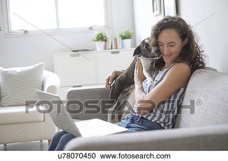 Stock Image - Young woman with laptop cuddling with her dog on sofa. Fotosearch