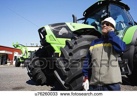 Stock Image - Farmer on the phone in front of tractor. Fotosearch