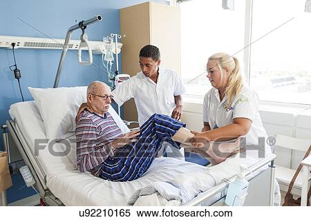 Stock Photography - Nurses helping senior man into hospital bed. Fotosearch
