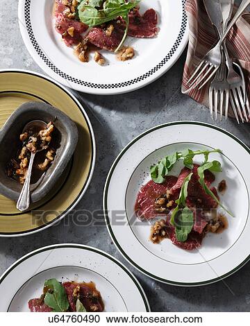 Stock Image - Bistro table with plates of beef carpaccio, rocket and walnuts. Fotosearch