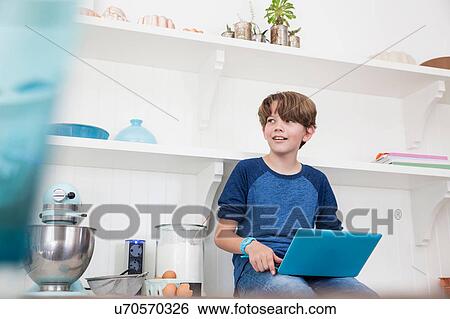 Stock Photograph - Boy sitting on kitchen work surface, using laptop computer. Fotosearch