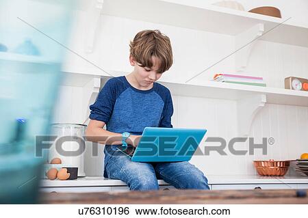 Stock Photograph - Boy sitting on kitchen work surface, using laptop computer. Fotosearch
