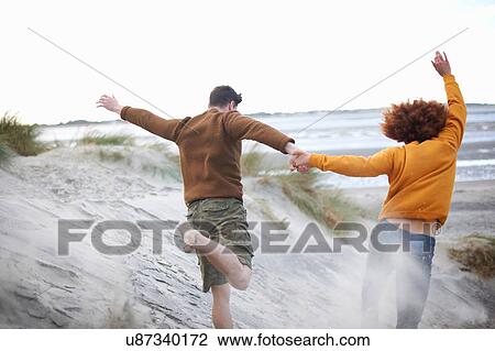 Stock Image - Couple running down sand dunes to beach. Fotosearch