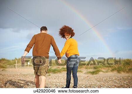 Stock Photograph - Couple walking on beach towards rainbow. Fotosearch