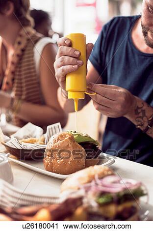 Stock Image - Cropped shot of couple putting mayonnaise on burger at sidewalk cafe. Fotosearch