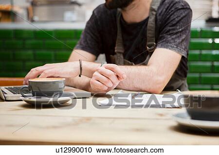 Stock Image - Cropped shot of male cafe owner doing paperwork at table. Fotosearch