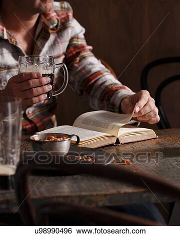 Stock Photograph - Cropped shot of man drinking beer and reading book at pub table. Fotosearch