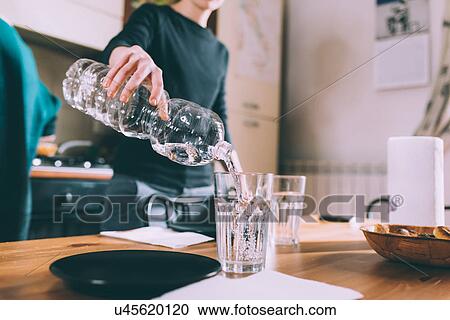 Stock Image - Cropped shot of young woman pouring water at kitchen table. Fotosearch