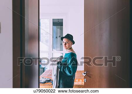 Stock Image - Doorway portrait of young woman wearing trilby standing in bedroom. Fotosearch