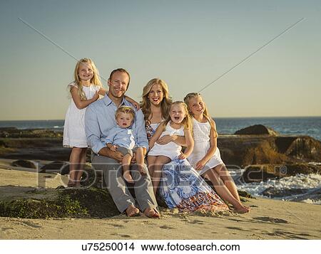 Picture - Family with children (12-17 months, 4-5, 6-7, 8-9) sitting on beach with sea in background. Fotosearch