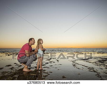 Stock Image - Father crouching next to daughter (4-5) on beach. Fotosearch