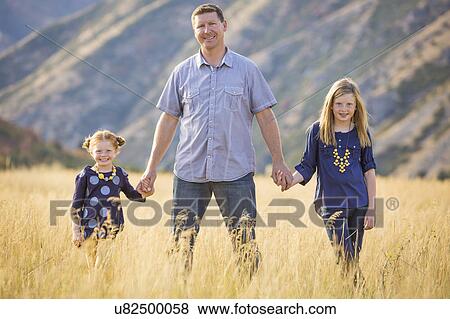 Stock Photo - Father with two daughters (4-5, 8-9) walking in field. Fotosearch