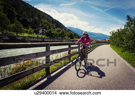 Picture - Female cyclist cycling along path by river in Vinschgau Valley, South Tyrol, Italy. Fotosearch
