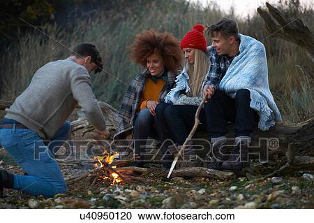 Stock Image - Four adult friends burning driftwood campfire on beach at dusk. Fotosearch
