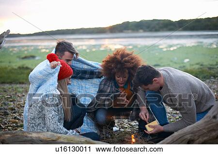 Stock Image - Four adult friends igniting campfire on beach. Fotosearch