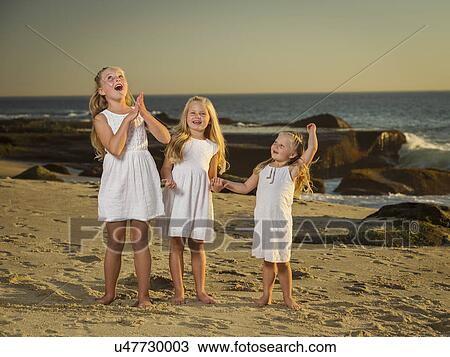 Stock Image - Girls (4-5, 6-7, 8-9) standing on beach with sea in background. Fotosearch