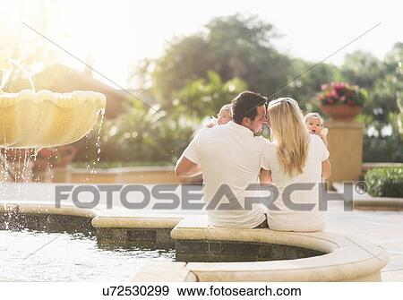 Stock Photo - Happy family with two baby girls (2-5 months) near fountain in sunlight. Fotosearch