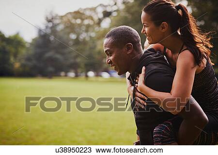 Stock Image - Man and woman having fun training in park, giving piggy back. Fotosearch