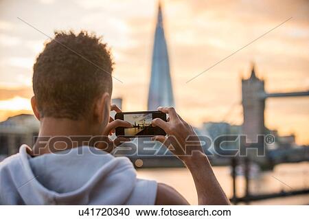 Stock Image - Man by riverside taking photograph of Tower Bridge and The Shard, Wapping, London, UK. Fotosearch