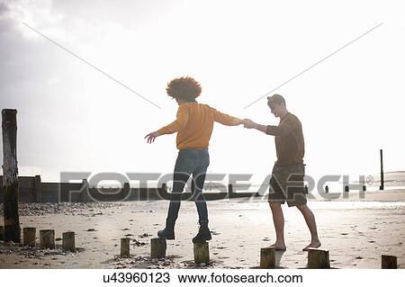 Stock Image - Man helping woman balance on wooden stumps on beach. Fotosearch