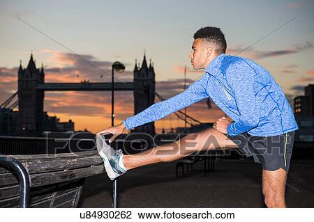 Stock Image - Man stretching by riverside, Tower Bridge in background, Wapping, London, UK. Fotosearch