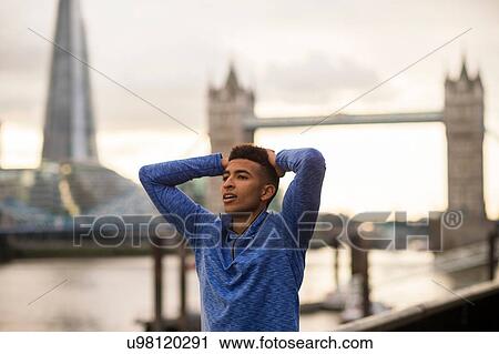 Stock Image - Man stretching by riverside, Tower Bridge and The Shard in background, Wapping, London, UK. Fotosearch