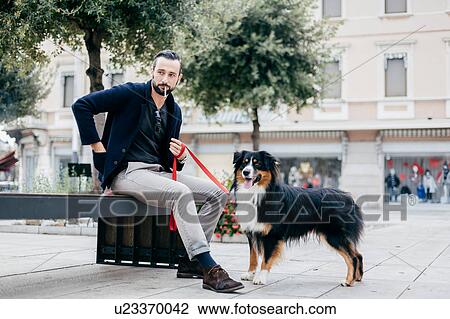 Stock Image - Man with dog smoking cigarette on city street bench. Fotosearch