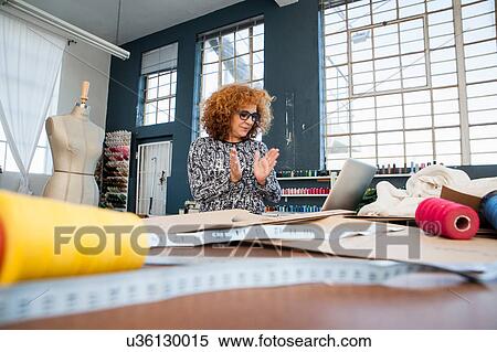 Stock Photography - Mature female fashion clapping at laptop on workshop table. Fotosearch