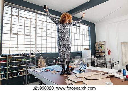 Stock Image - Mature female fashion designer dancing to smartphone music on workshop table. Fotosearch