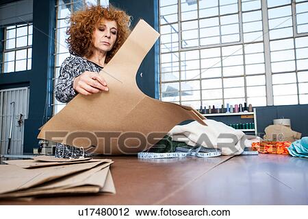 Stock Image - Mature female fashion designer preparing pattern on workshop table. Fotosearch
