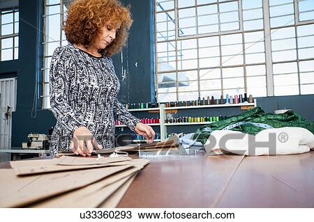 Stock Image - Mature female fashion designer preparing pattern on workshop table. Fotosearch
